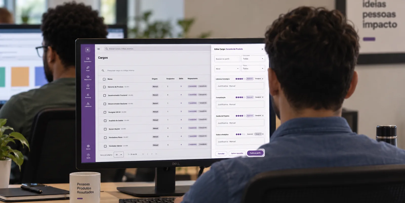 Foto de um homem de camisa azul visto de costas, diante de um monitor com a tabela “Cargos” e um painel lateral de edição de perfil, com caneca, caderno e escritório desfocado ao fundo.