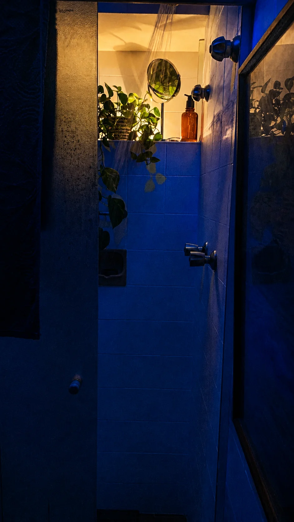 Box de banheiro interno visto da porta, com espelho redondo, plantas e frasco âmbar sob luz quente contra azulejo azulado